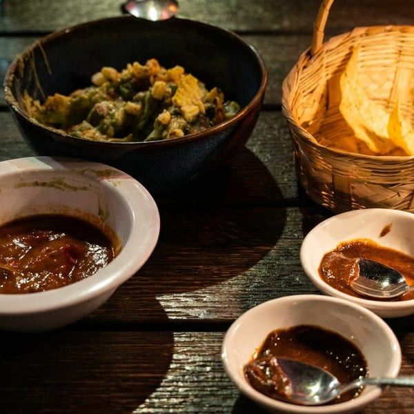 Guacamole, tortilla chips in a basket, and three bowls of salsa on a wooden table.