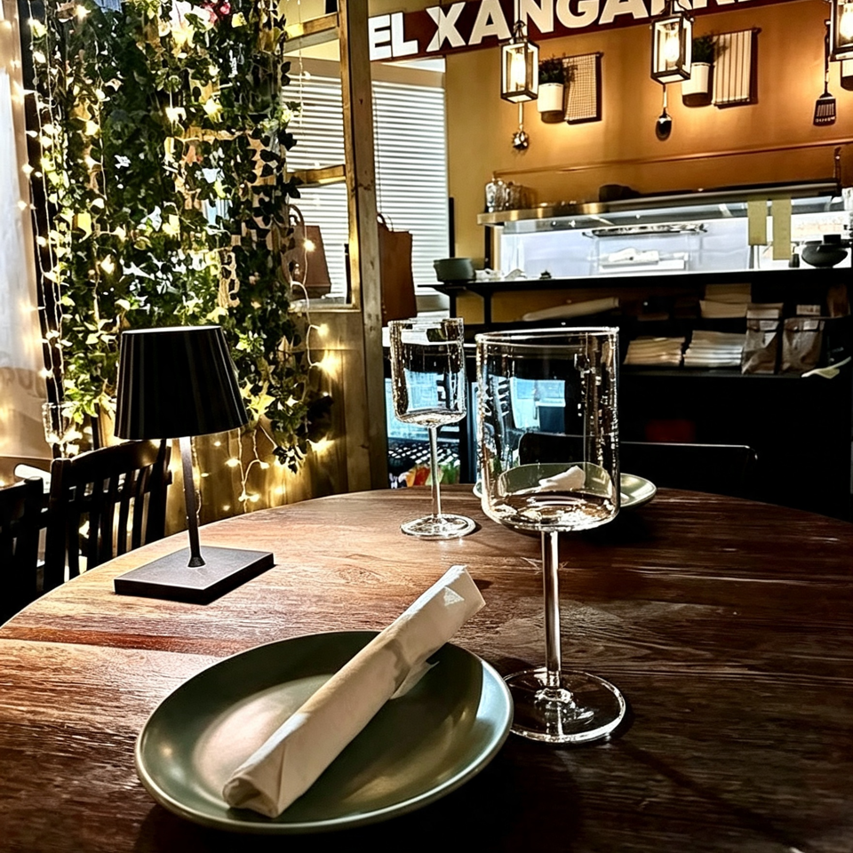 Warmly lit El Xangarrito restaurant interior with a set wooden table, wine glasses, greenery, and illuminated bar.