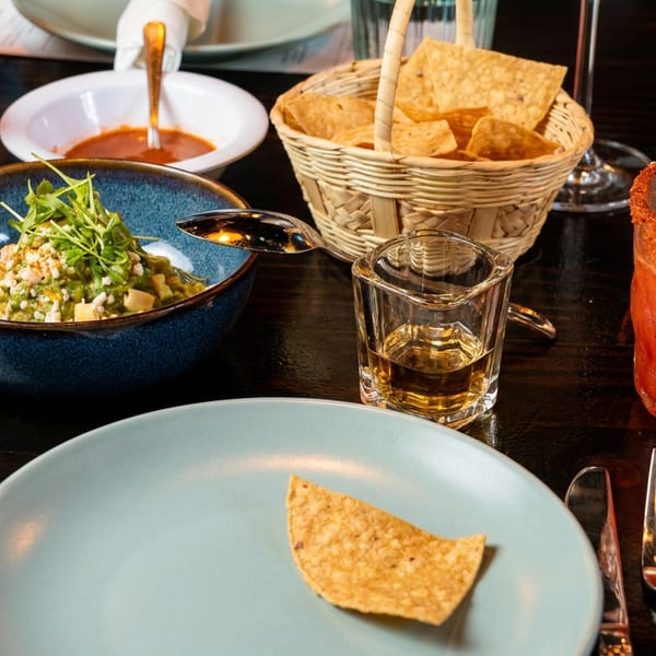 A table set with a blue bowl of ceviche, tortilla chips, red drinks, and a shot of tequila.