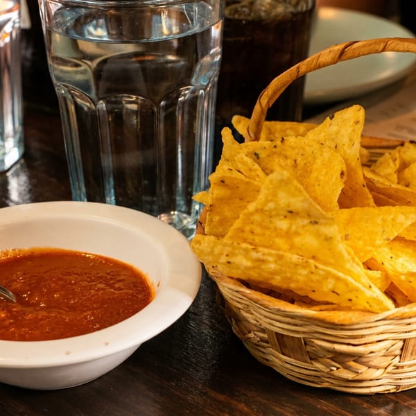 A basket of tortilla chips and a bowl of salsa with a spoon on a dark wooden table.