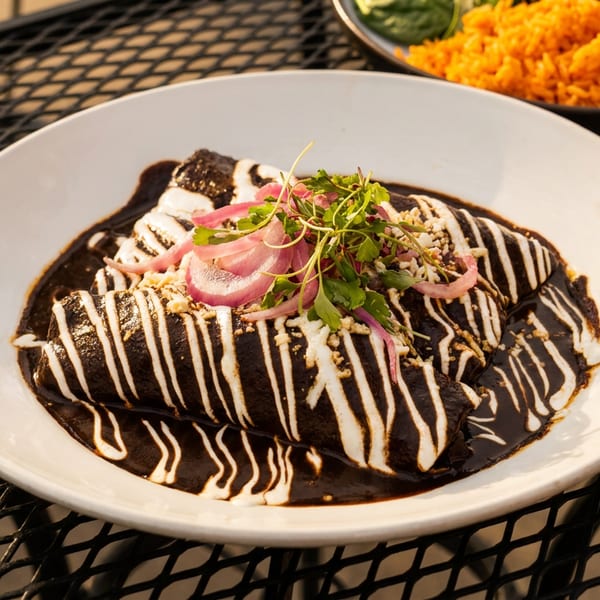 Enchiladas with dark mole sauce, white crema, red onions, and microgreens on a white plate.