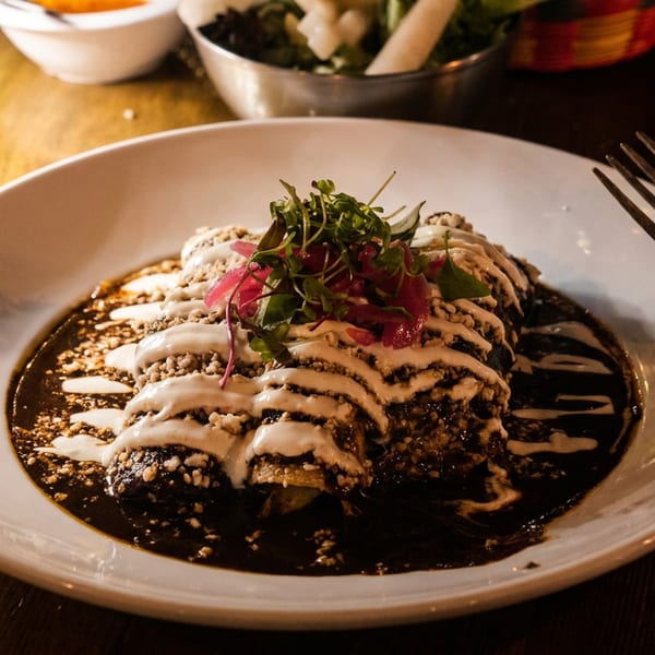 Enchiladas with dark mole sauce, white crema, crumbled cheese, and microgreens on a white plate.