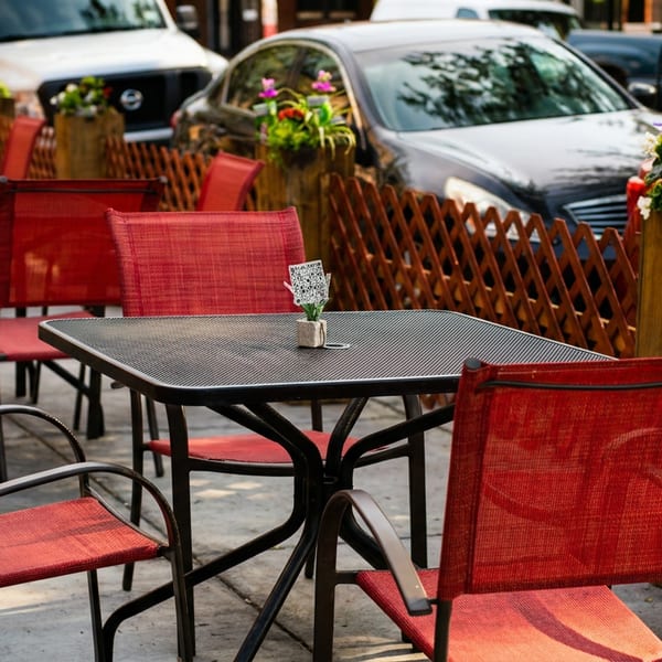 Outdoor patio dining area with red chairs, black tables, and potted plants, cars parked in background.