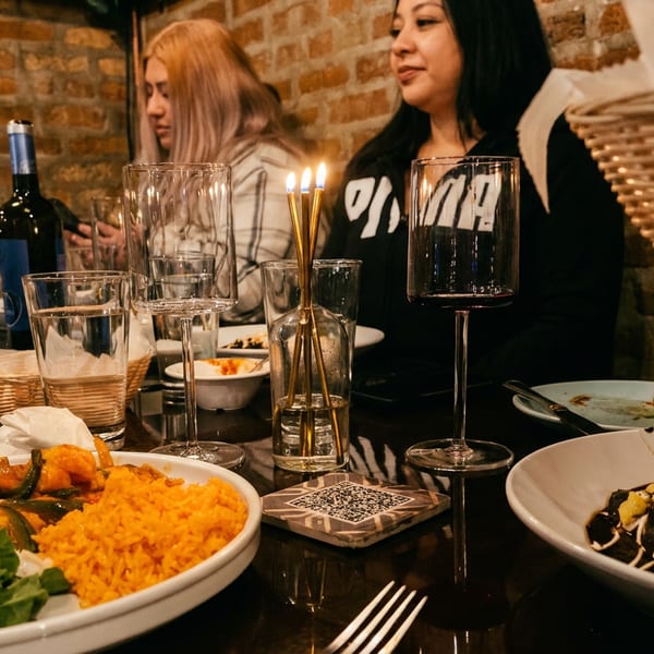 Diners at a restaurant table with plates of food, wine, and a lit candle against a brick wall.
